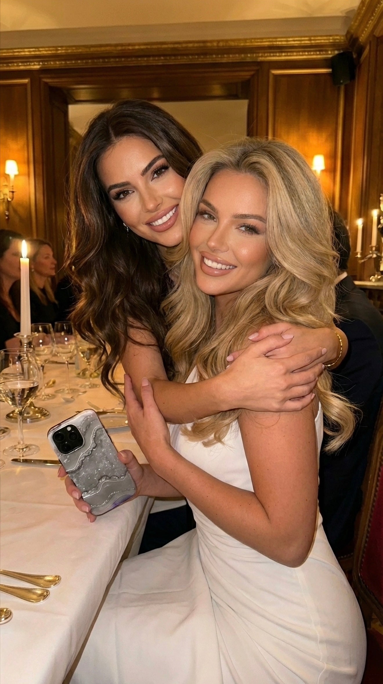 Two smiling women in elegant attire hug at a stylish dinner setting, one holding a trendy Silver Marble Quartz phone case with sølv, gråt, and flerfarvet tones.