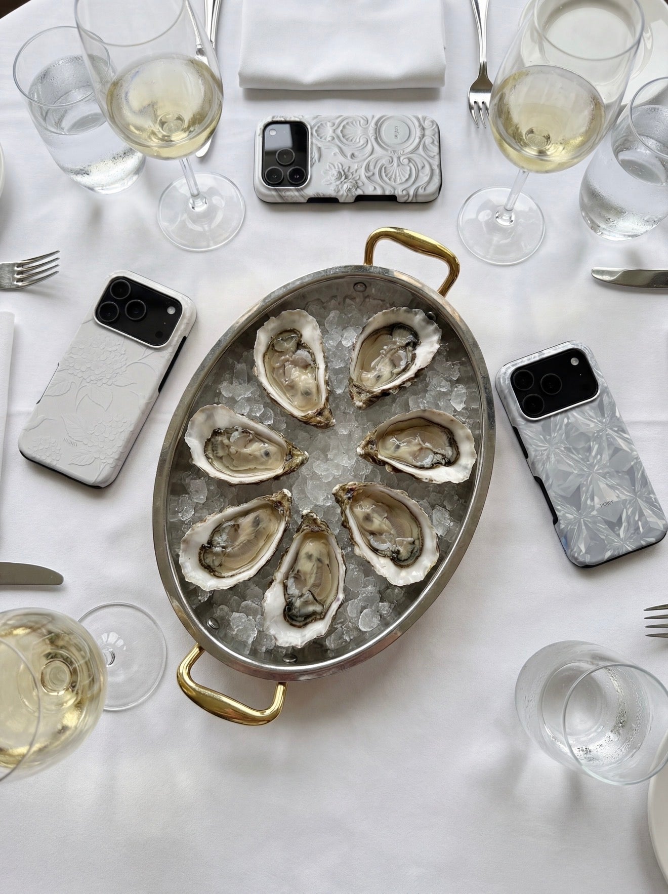 A stylish table setting with white wine glasses, oysters on ice in a silver dish, and trendy phone cases in flerfarvet, hvid, and beige tones.