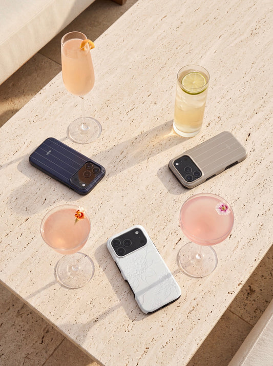Three stylish phone cases in flerfarvet and hvid colors placed on a beige table surrounded by elegant pink and light-colored cocktails.