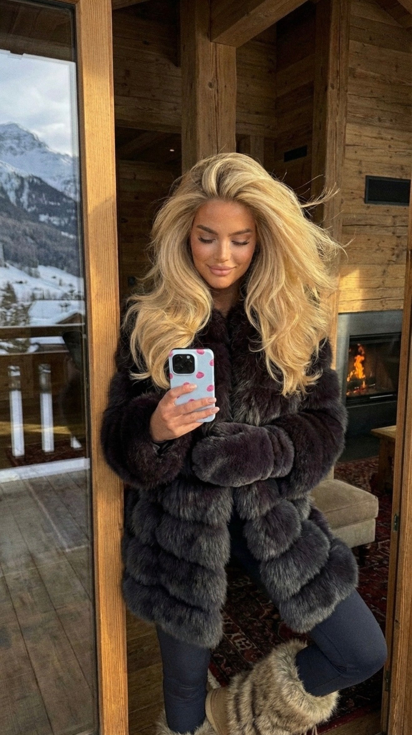 Trendy young woman with voluminous blonde hair taking a selfie inside a cozy cabin wearing stylish winter attire and a phone case with a raspberry pattern.