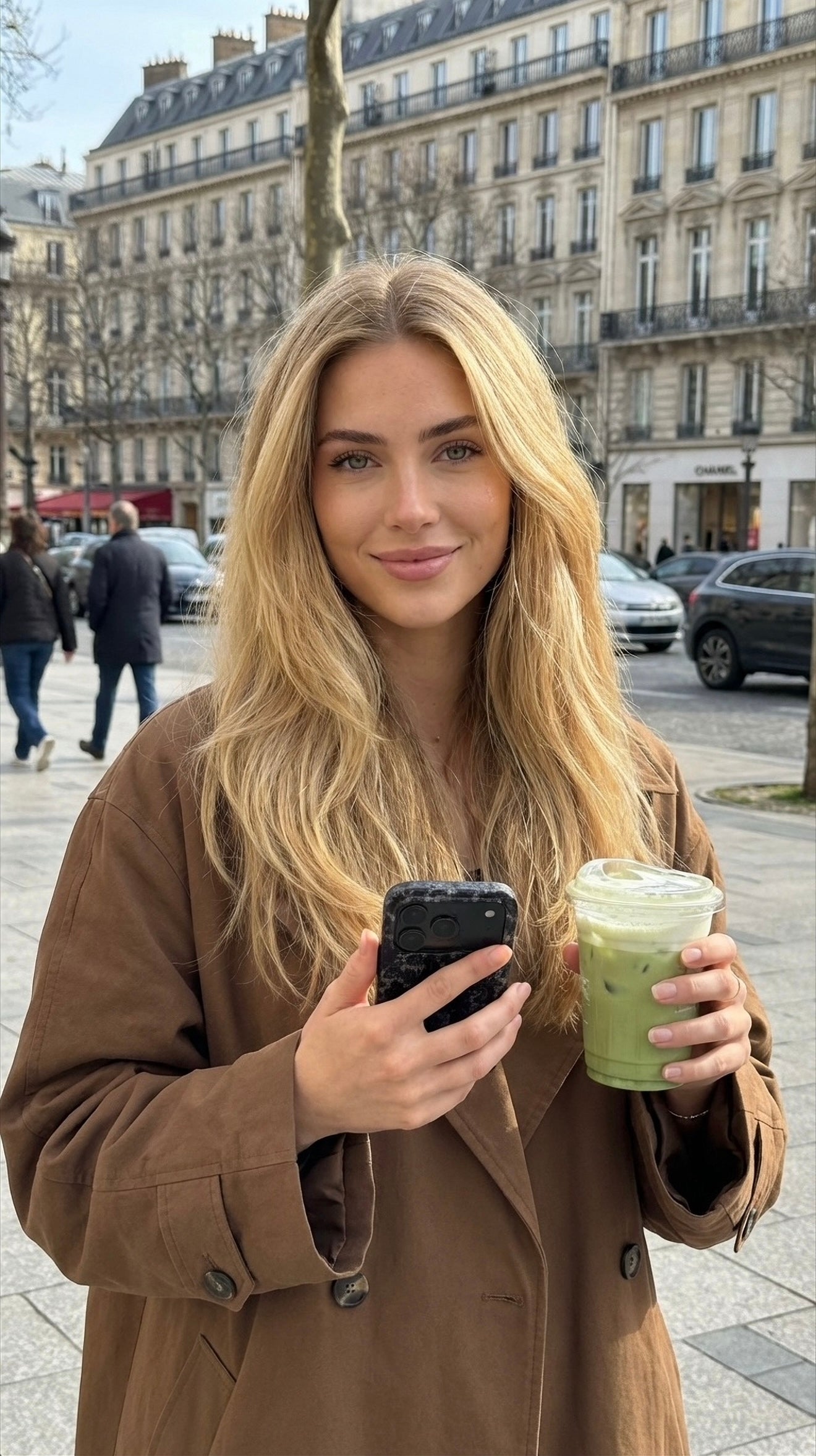 Trendy young woman with long blonde hair holding a drink and phone with stylish black phone case, standing in an urban outdoor setting with elegant background.