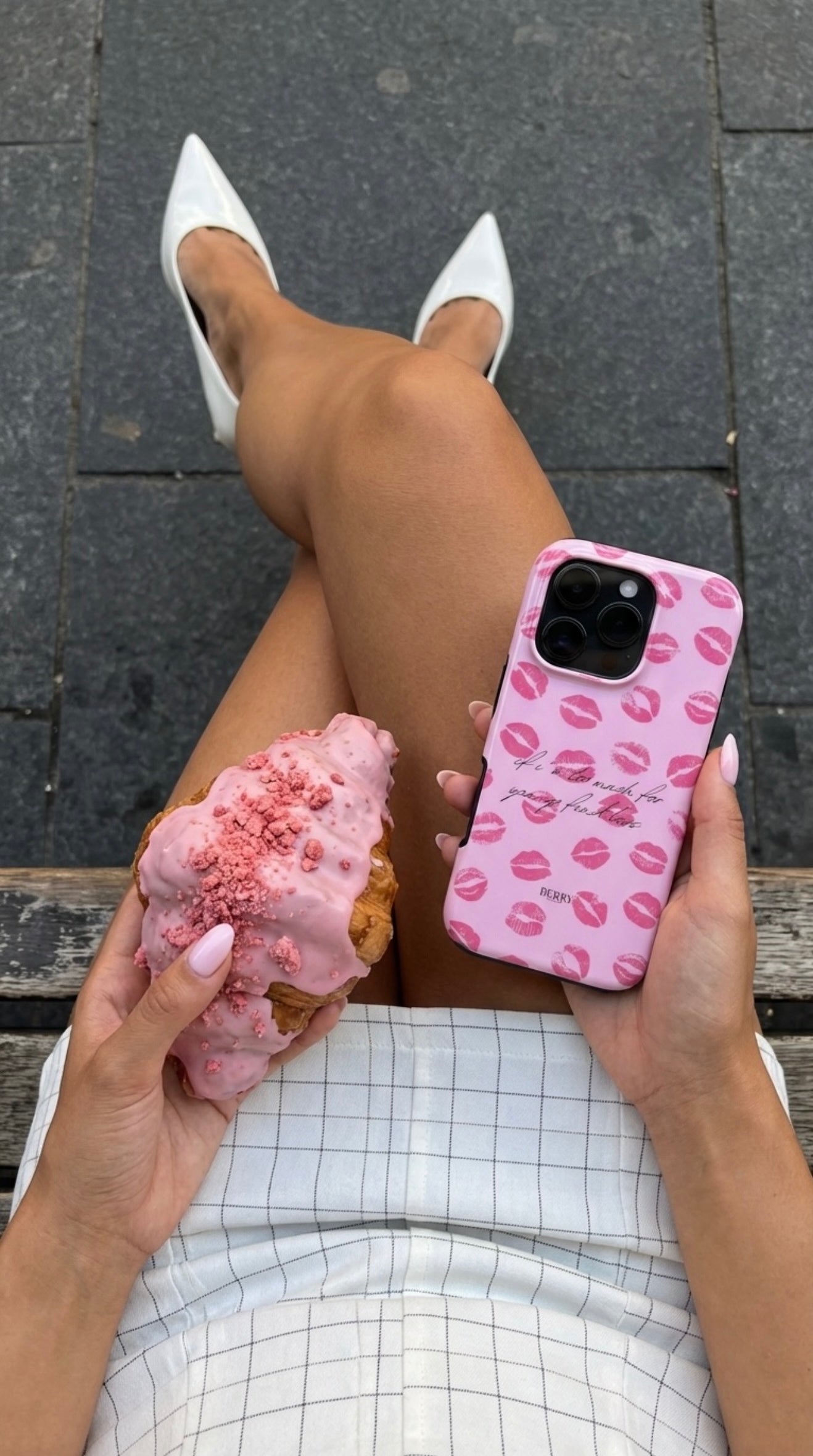 Trendy pink phone case with lips pattern held by a feminine hand with matching nails, paired with a stylish pink iced pastry and white pointed heels.