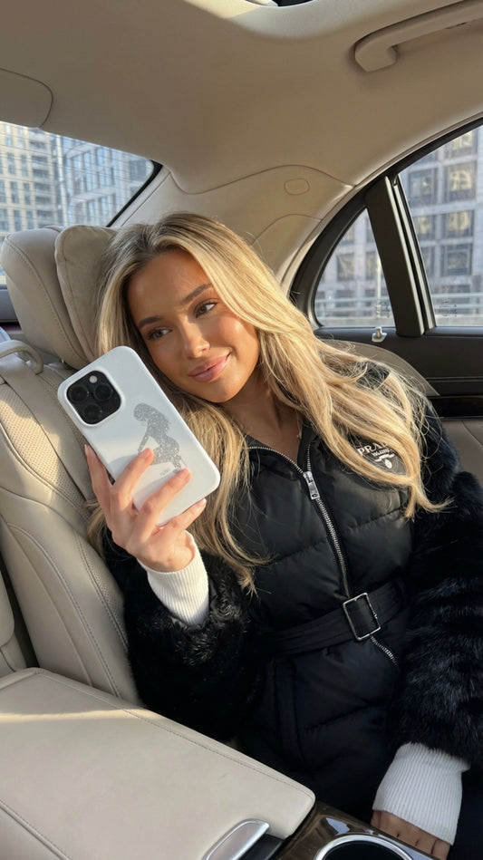 Trendy girl with blonde hair sitting in car holding phone with white and multicolored phone case featuring silver and gray accents, smiling and looking at screen.