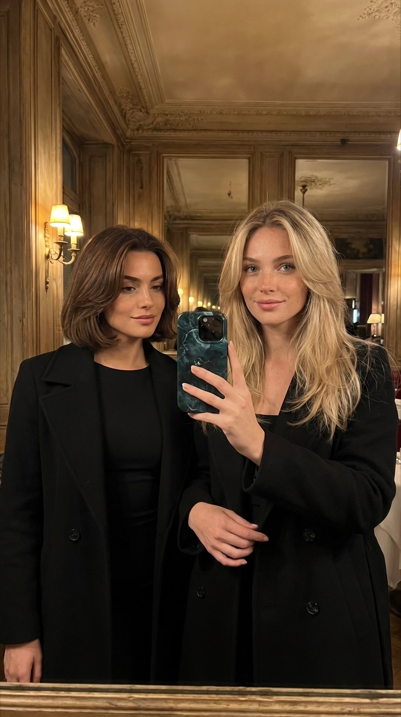 Two stylish women wearing black coats taking a mirror selfie with a trendy green patterned phone case in an elegant vintage room.