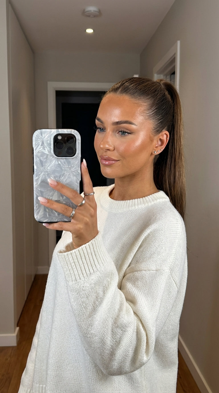 Trendy woman with sleek ponytail wearing a white sweater holding a phone with a stylish silver-white multicolored phone case reflecting light beautifully.