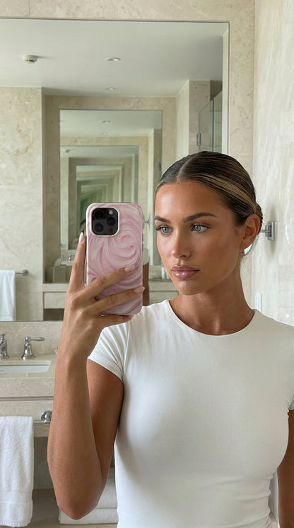 Trendy woman taking a mirror selfie holding a phone with a pink, flerfarvet creamy strawberry swirl patterned case, showcasing a feminine and stylish look.