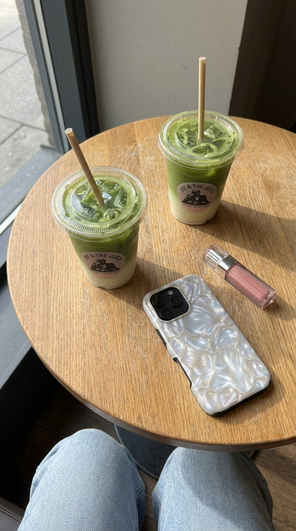 Trendy phone case with a magical silver pearlescent pattern on a wooden table, next to two green layered drinks and a soft pink lip gloss for a feminine vibe.