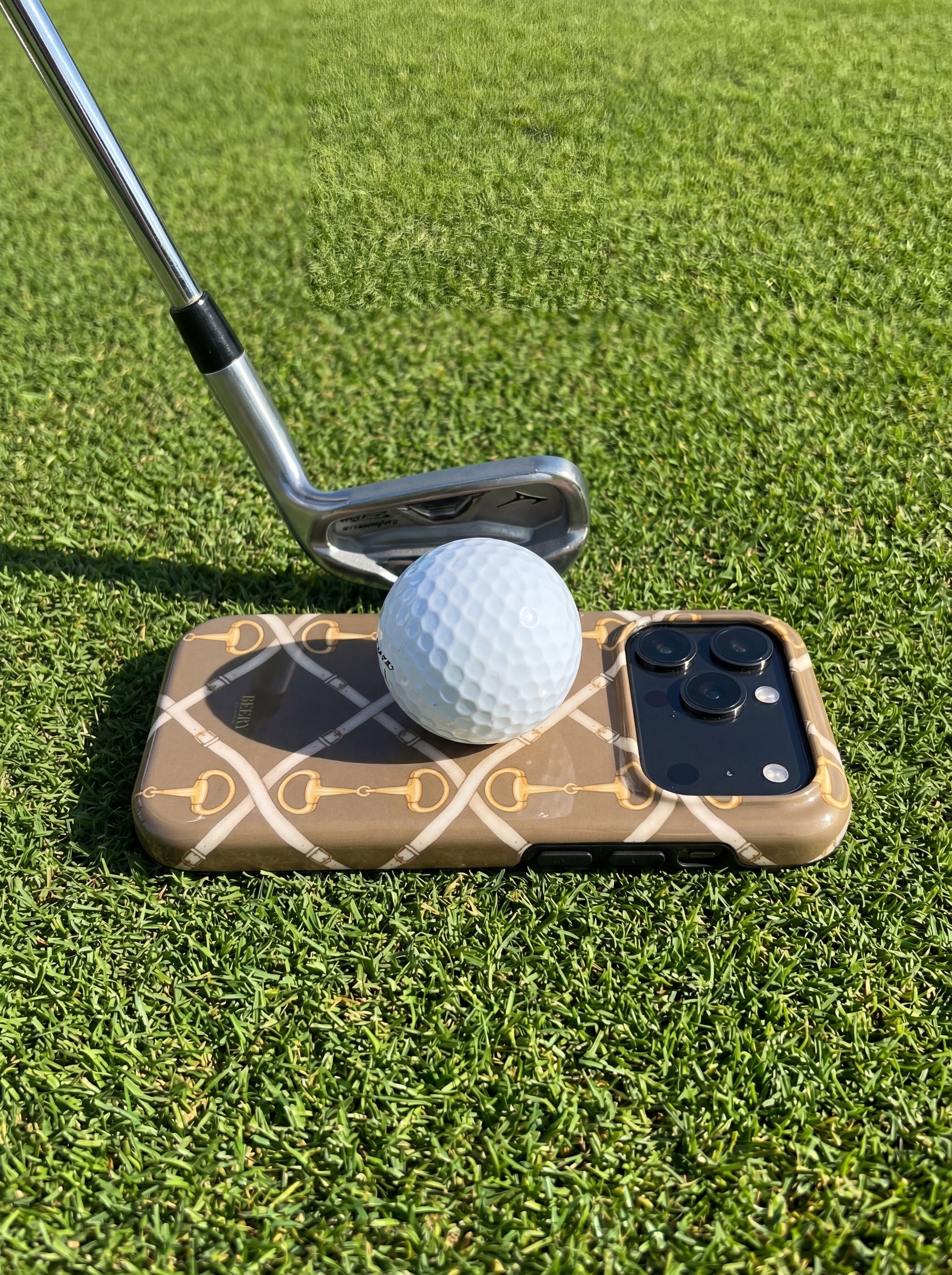 Trendy phone case with brown, beige, and gold buckle pattern under a golf ball and club on vibrant green grass, reflecting a stylish feminine aesthetic.