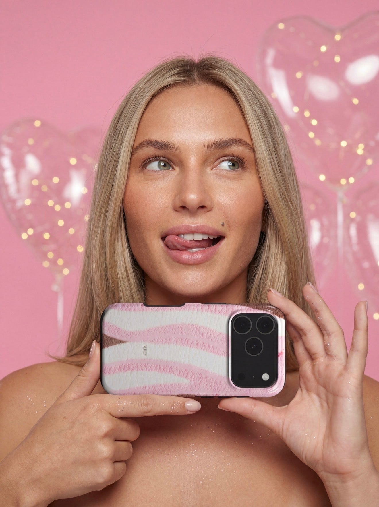 Young woman holding a trendy pink and white patterned phone case with a playful expression against a pink background with heart-shaped balloons.