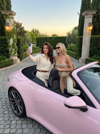 Two stylish women in neutral tones taking a selfie in a pink convertible on a cobblestone driveway with green hedges and lamp posts in the background.