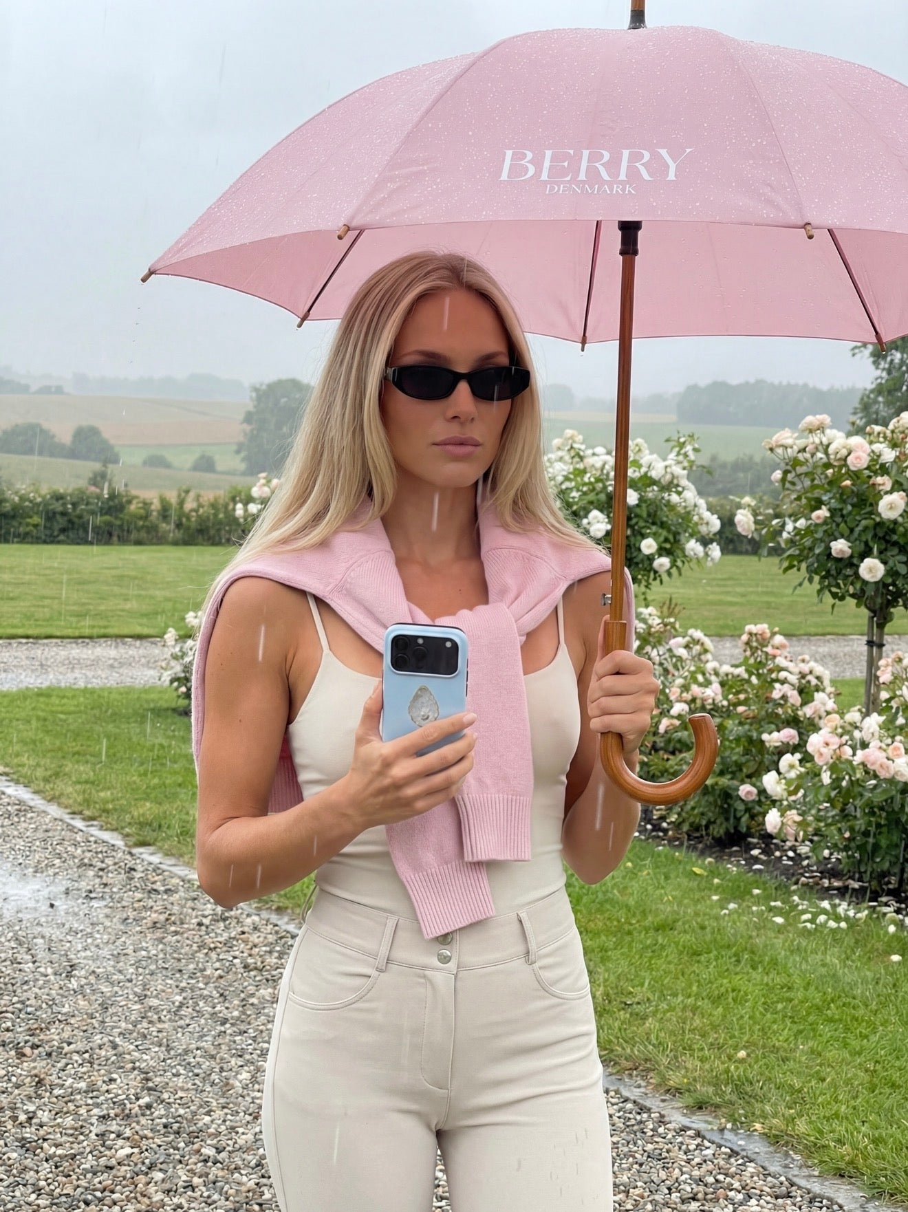 Trendy young woman holding a phone with a stylish blå, beige, flerfarvet, and guld phone case under a pink umbrella on a rainy day outdoors.
