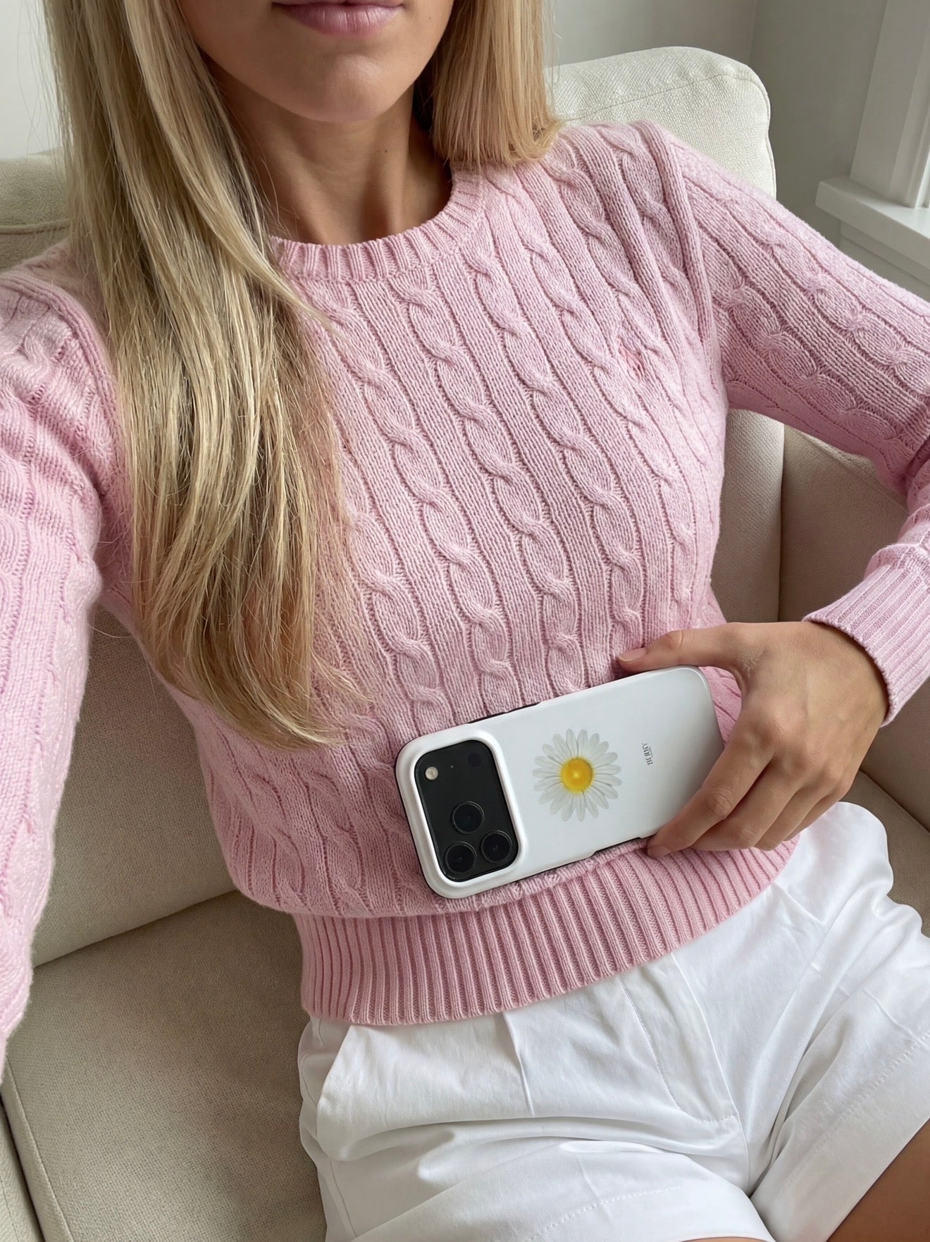 Woman in pink cable knit sweater holding phone with white daisy patterned phone case featuring yellow center and green accents