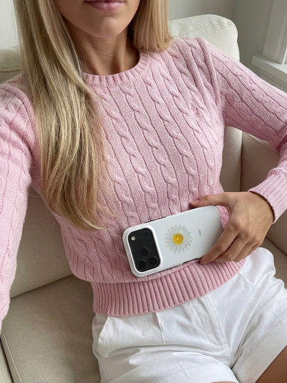 Woman in pink cable knit sweater holding phone with white daisy patterned phone case featuring yellow center and green accents