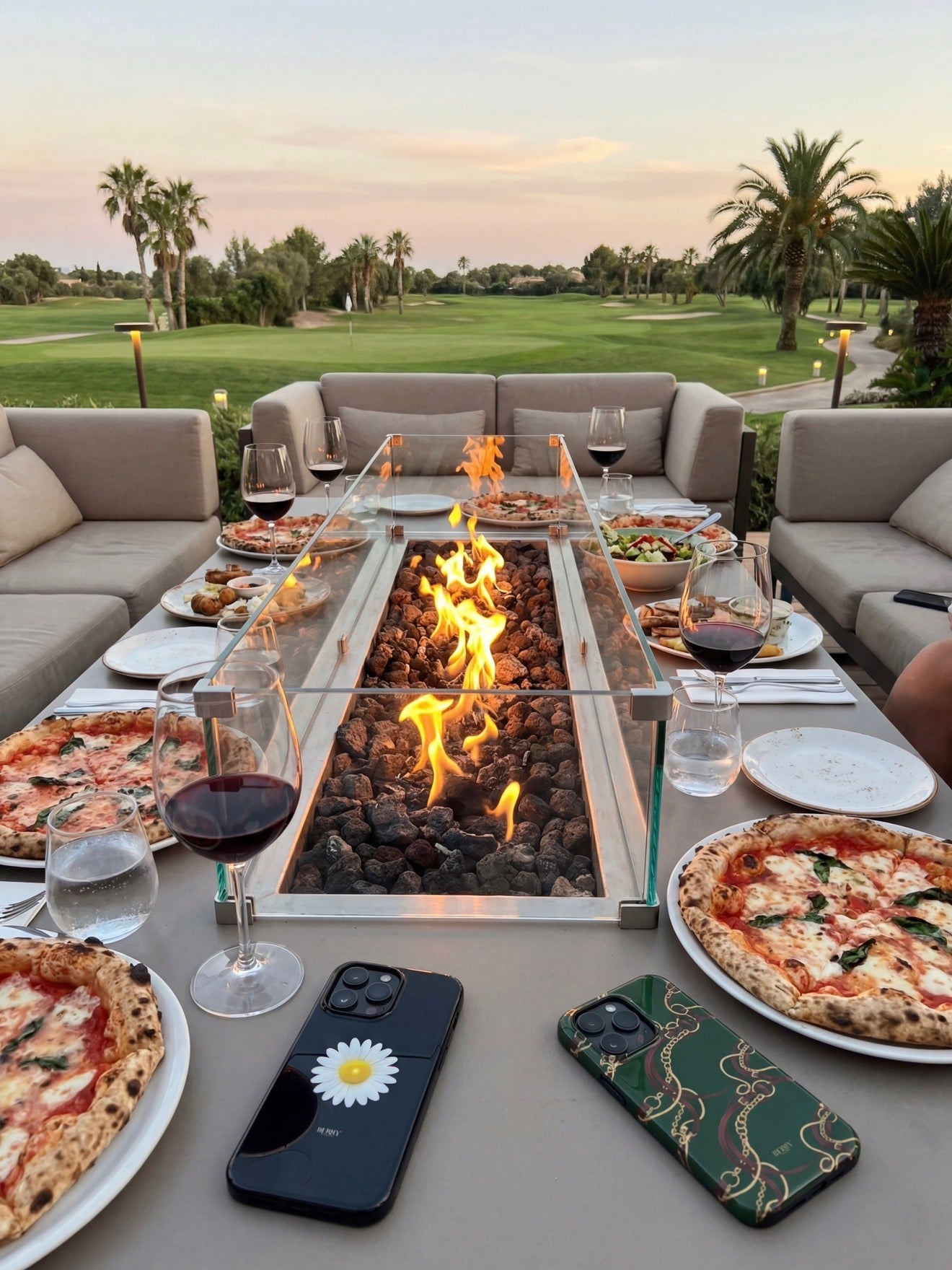 Outdoor seating area with a modern fire pit table surrounded by pizzas, glasses of red wine, and two stylish phone cases with trendy feminine designs.