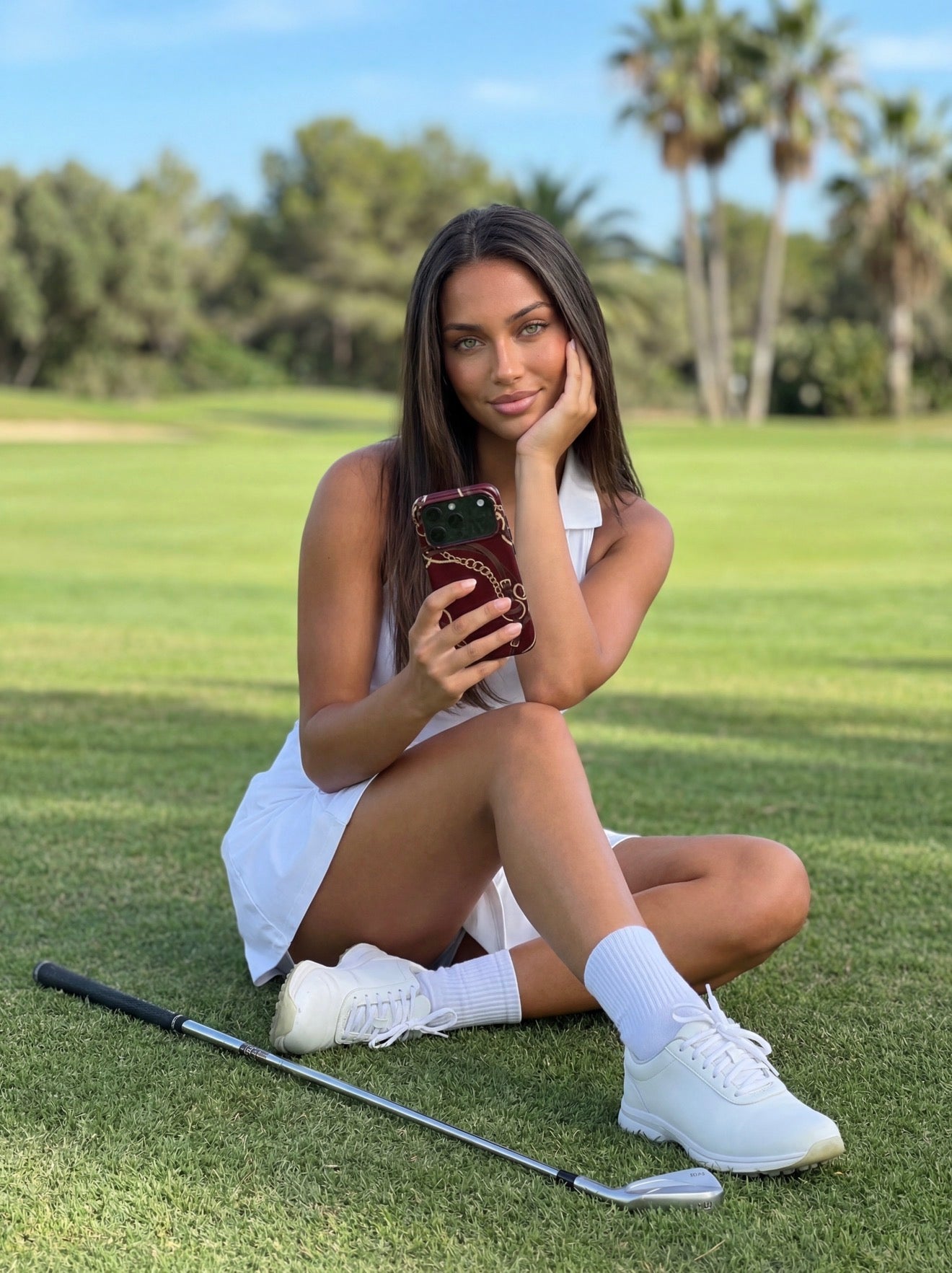 Trendy young woman sitting on grass outdoors holding a phone with a multicolored burgundy, gold, brown, beige, and red case, exuding chic feminine style.