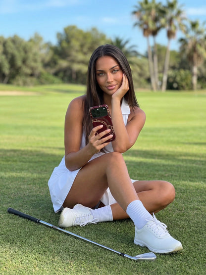 Trendy young woman sitting on grass outdoors holding a phone with a multicolored burgundy, gold, brown, beige, and red case, exuding chic feminine style.