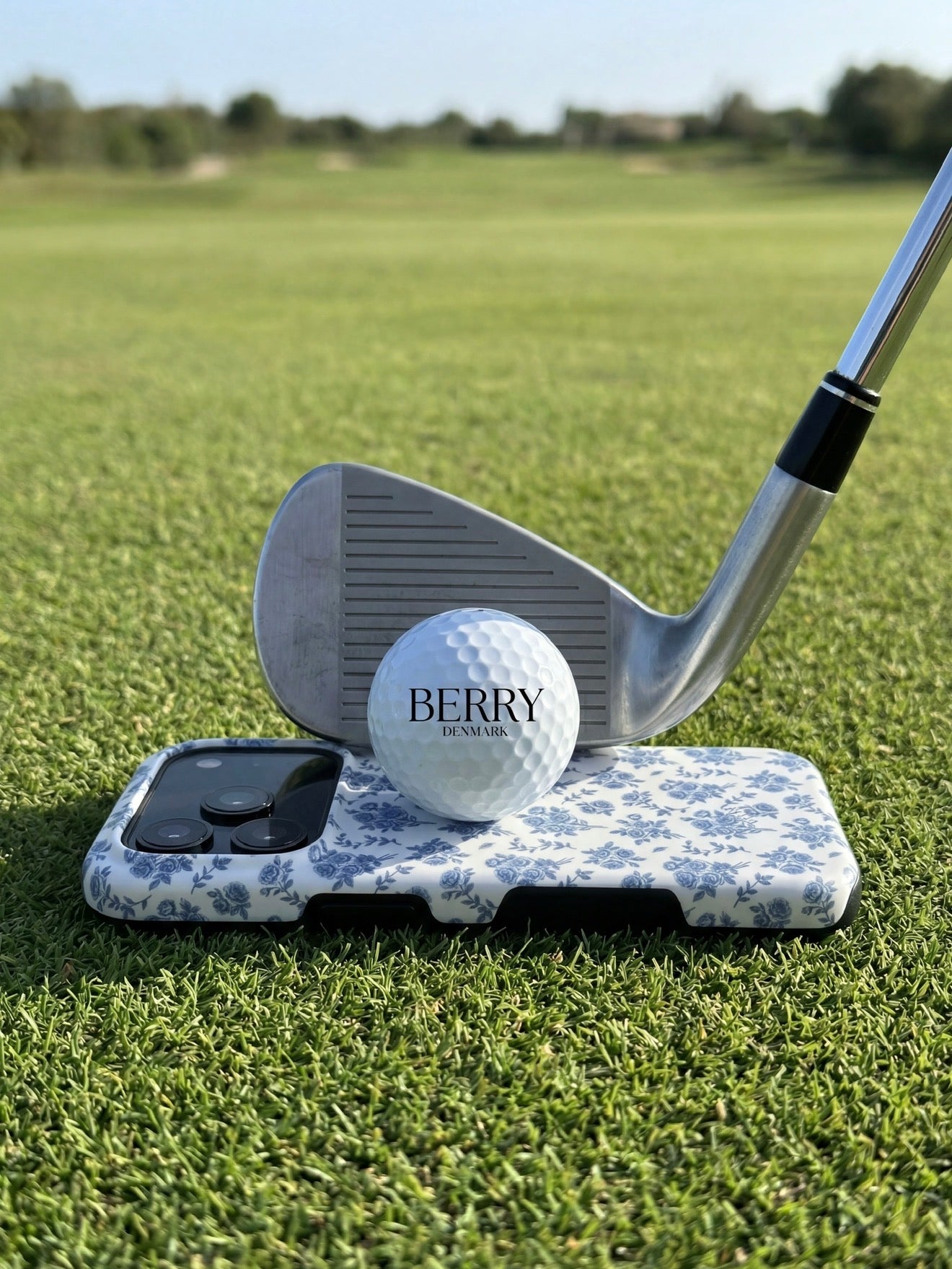 Trendy phone case with white and blue floral pattern placed on grass, with golf club and branded golf ball creating a stylish and feminine outdoor vibe.