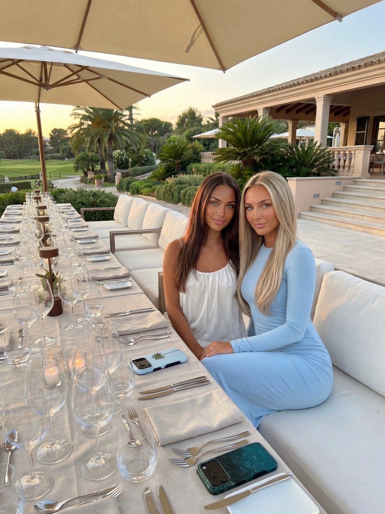 Two stylish women with feminine trendy outfits seated at an elegant outdoor dining table with phone cases in blue and green shades nearby.