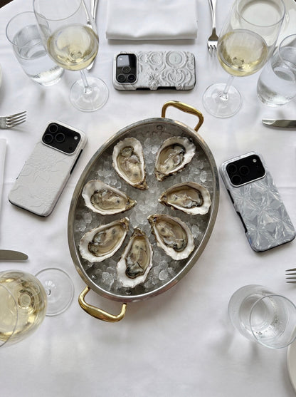 A stylish table setting with white wine glasses, oysters on ice in a silver dish, and trendy phone cases in flerfarvet, hvid, and beige tones.
