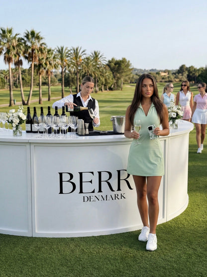 Trendy young woman in a feminine white and green outfit holding a glass and a flerfarvet, hvid, grøn phone case at an elegant outdoor event.