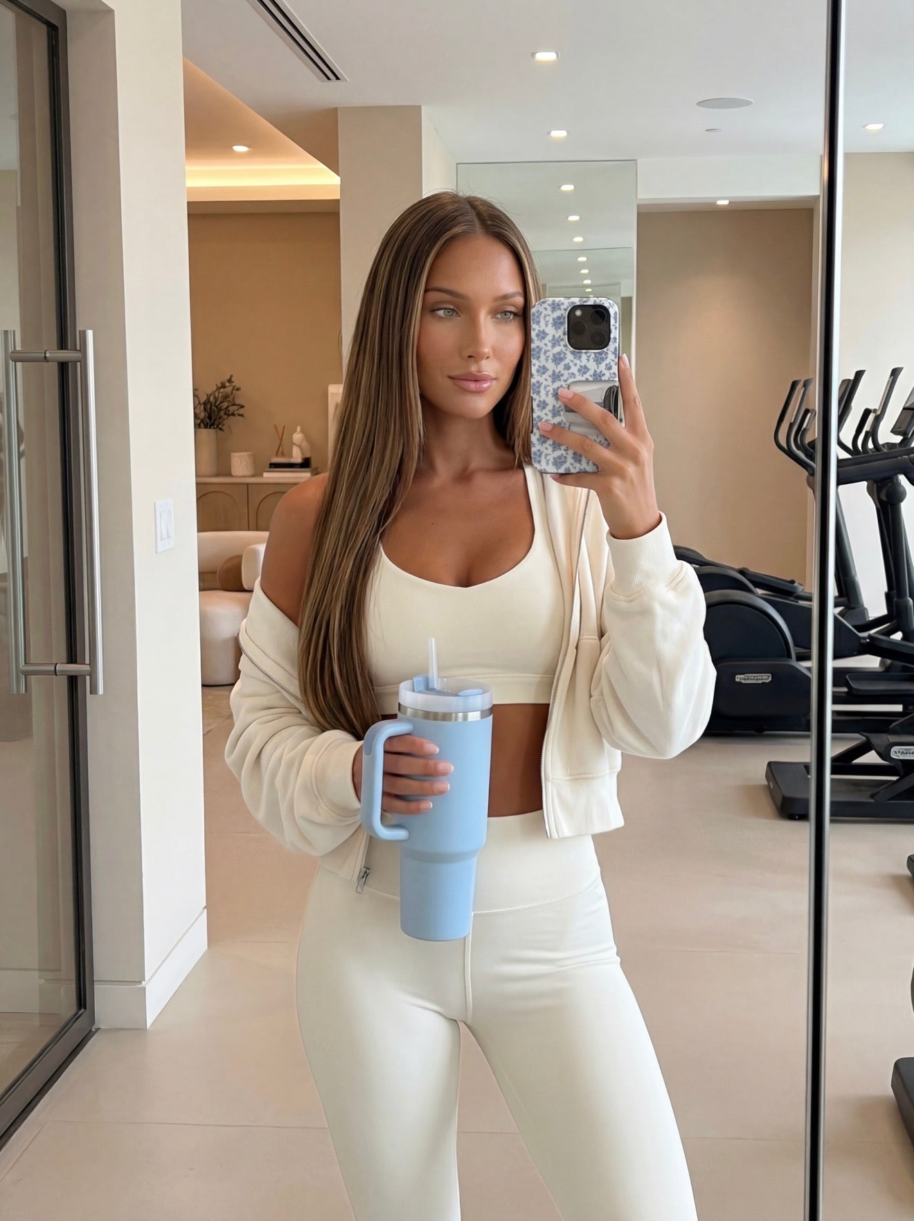 Trendy young woman taking a mirror selfie holding a blue and multicolored patterned phone case and a blue tumbler in a sleek, modern gym setting.