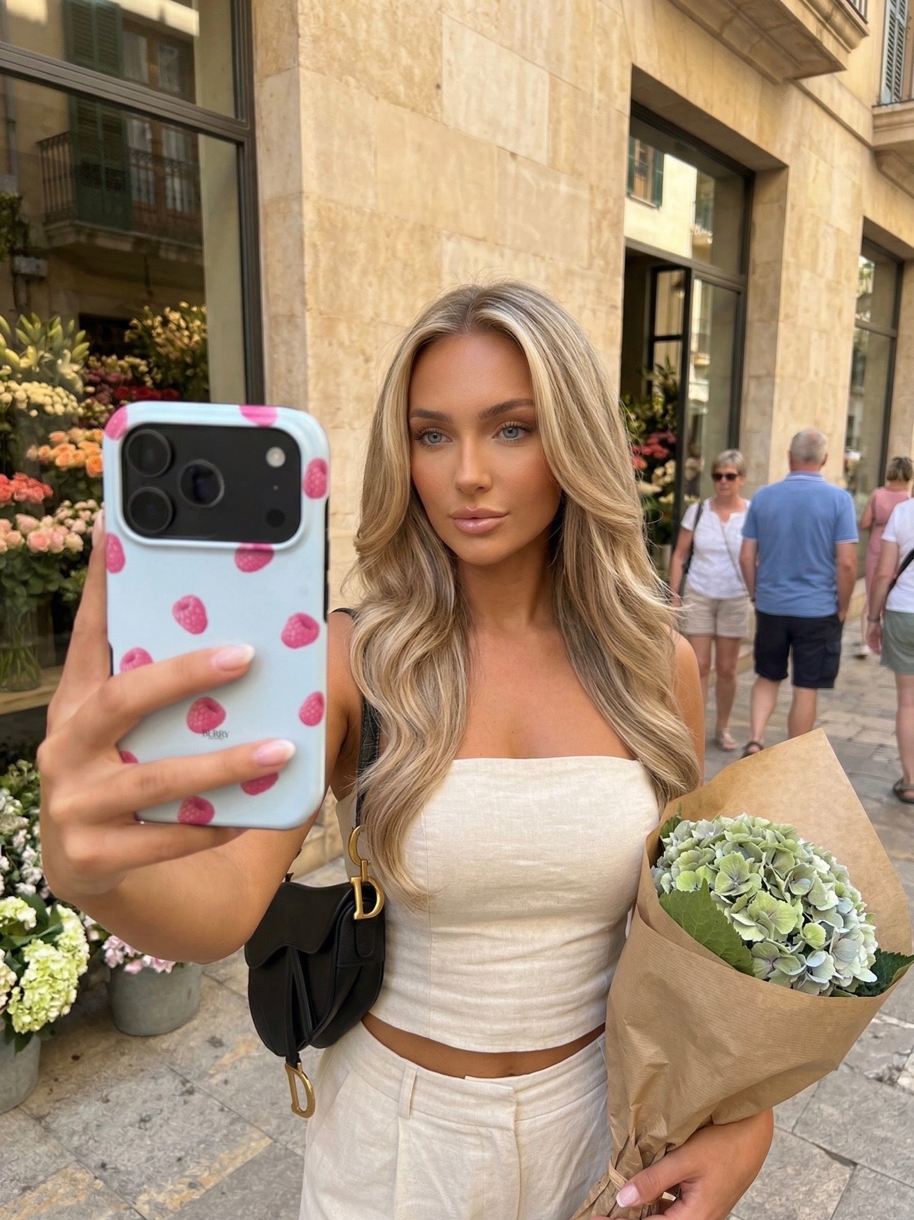 Trendy girl taking a selfie outdoors holding a stylish phone case with raspberry pattern and a bouquet of flowers, exuding a feminine and beautiful vibe.