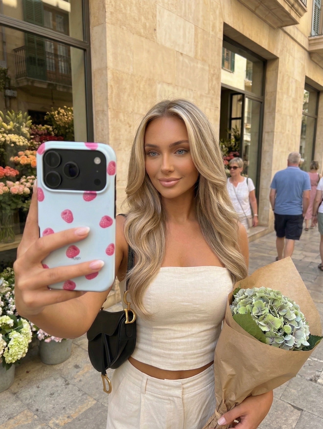 Trendy young woman with long blonde hair taking a selfie outdoors holding a bouquet, featuring a stylish phone case with playful raspberry design.