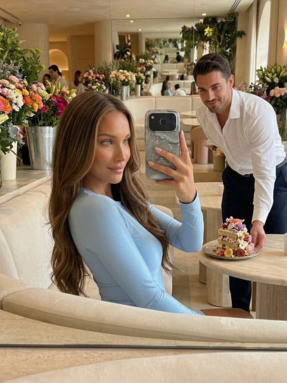 Woman with long hair takes a trendy selfie using a phone case with silver marble quartz pattern in a bright cafe with flowers and a man serving cake.