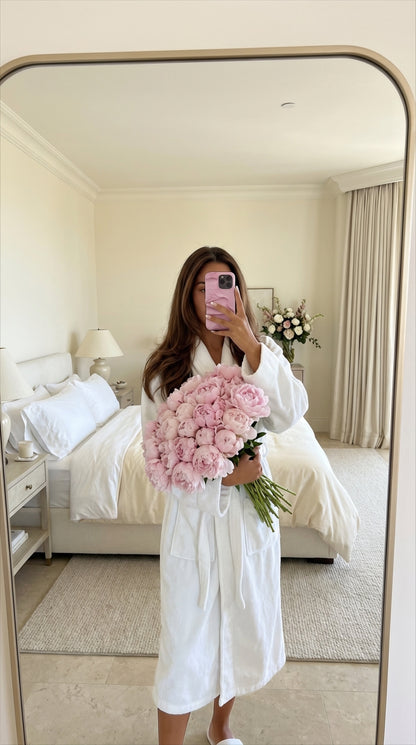 Trendy woman in white robe holding pink flerfarvet flowers with a pink phone case, in a bright feminine bedroom setting.