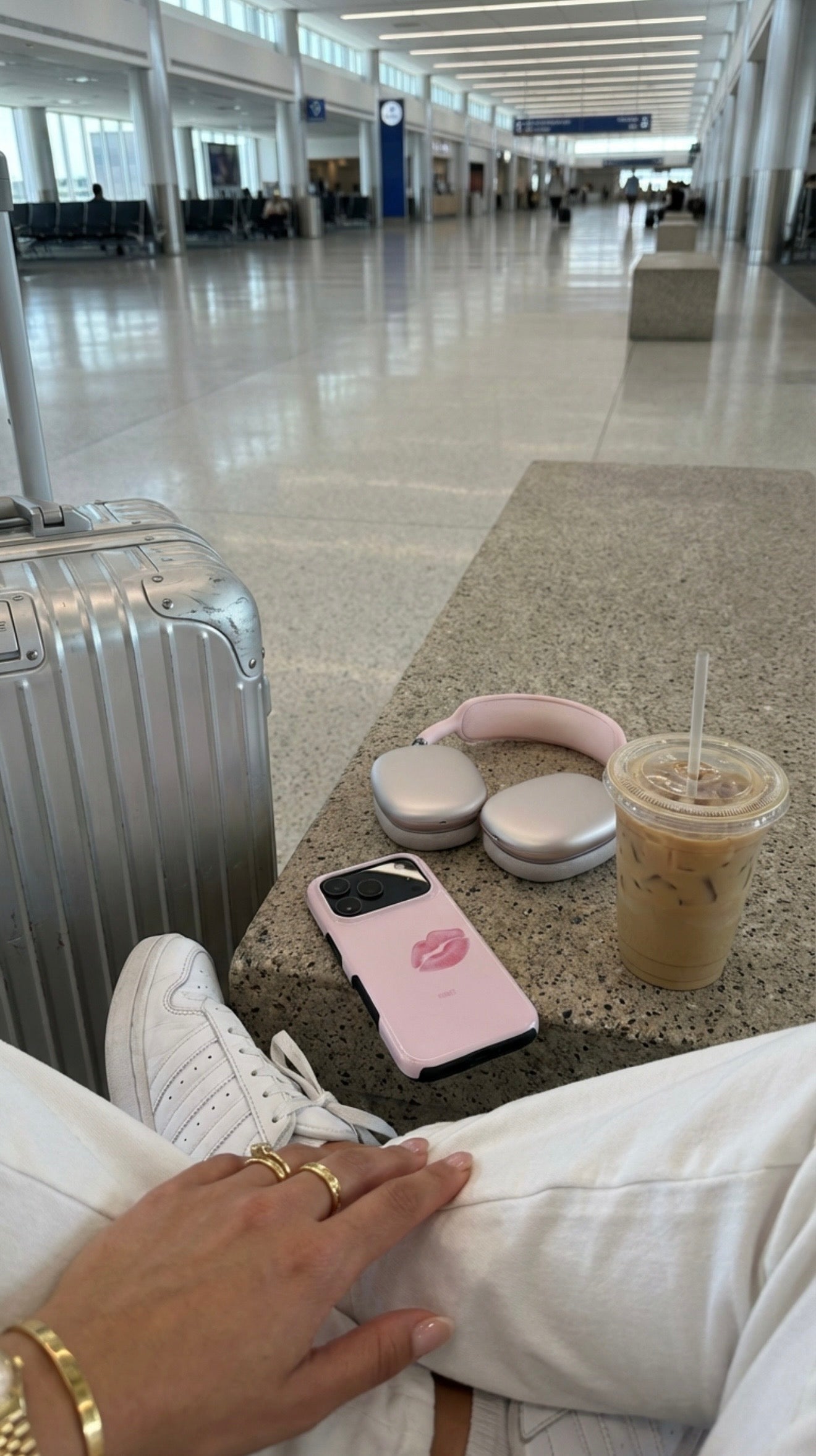Trendy feminine pink phone case with lips design, paired with matching pink headphones, styled with white sneakers and iced coffee in a modern travel setting.
