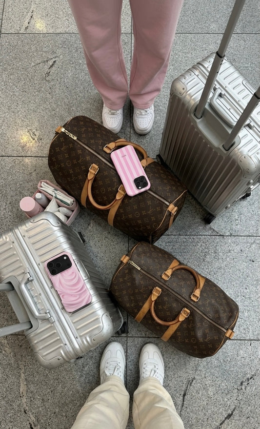 Two people standing with pink and white sneakers surrounded by silver suitcases, brown patterned bags, and pink stylish phone cases on airport floor.