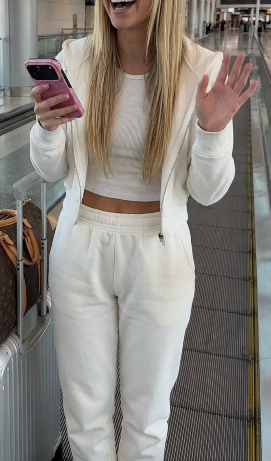 Trendy woman wearing casual white outfit holding phone with pink glitter case, smiling and waving on an airport moving walkway.