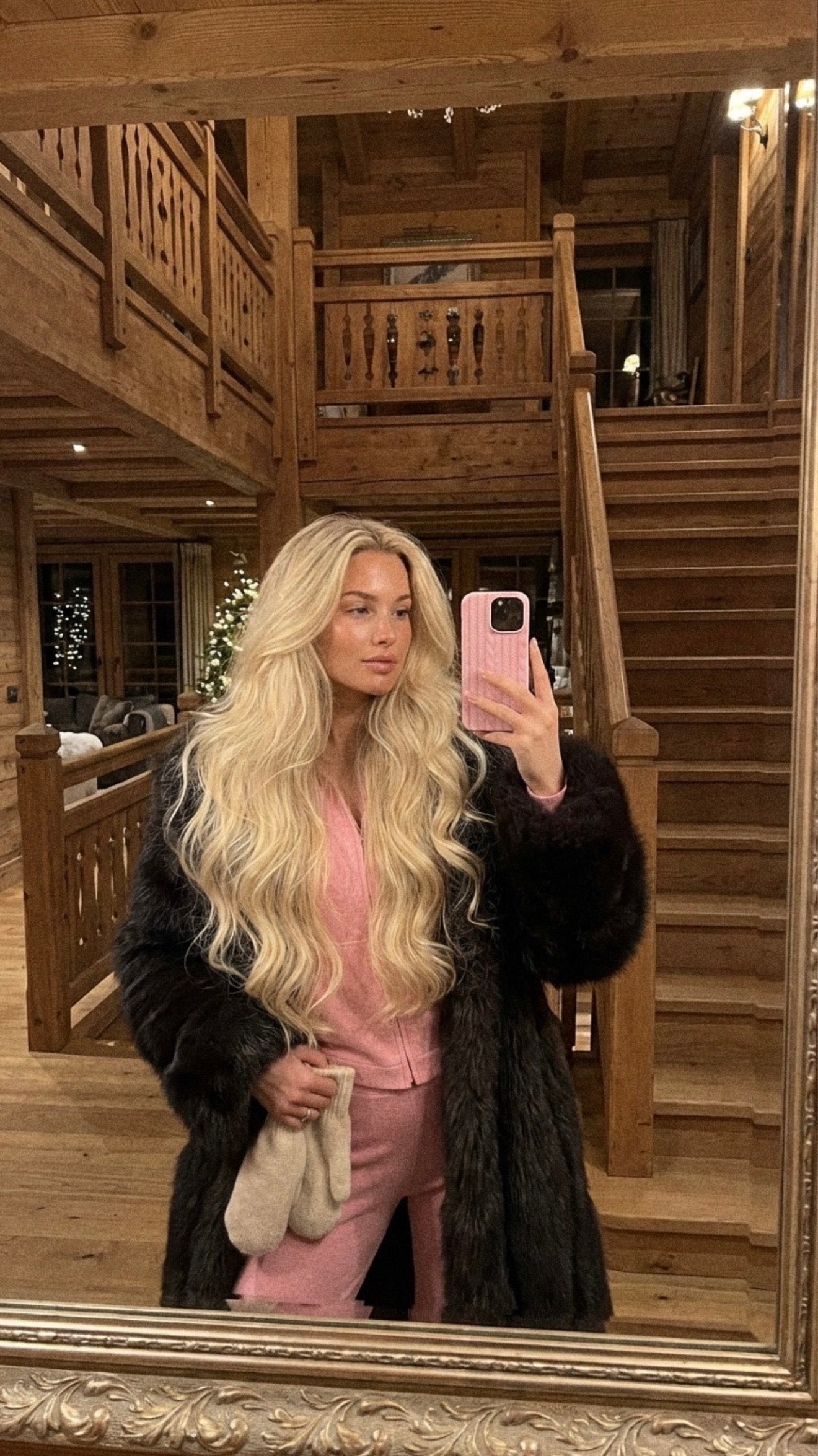 Trendy feminine woman with long blonde hair wearing a pink outfit and holding a phone with a pink flerfarvet case in a cozy wooden interior.
