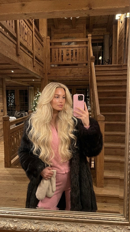 Trendy feminine woman with long blonde hair wearing a pink outfit and holding a phone with a pink flerfarvet case in a cozy wooden interior.