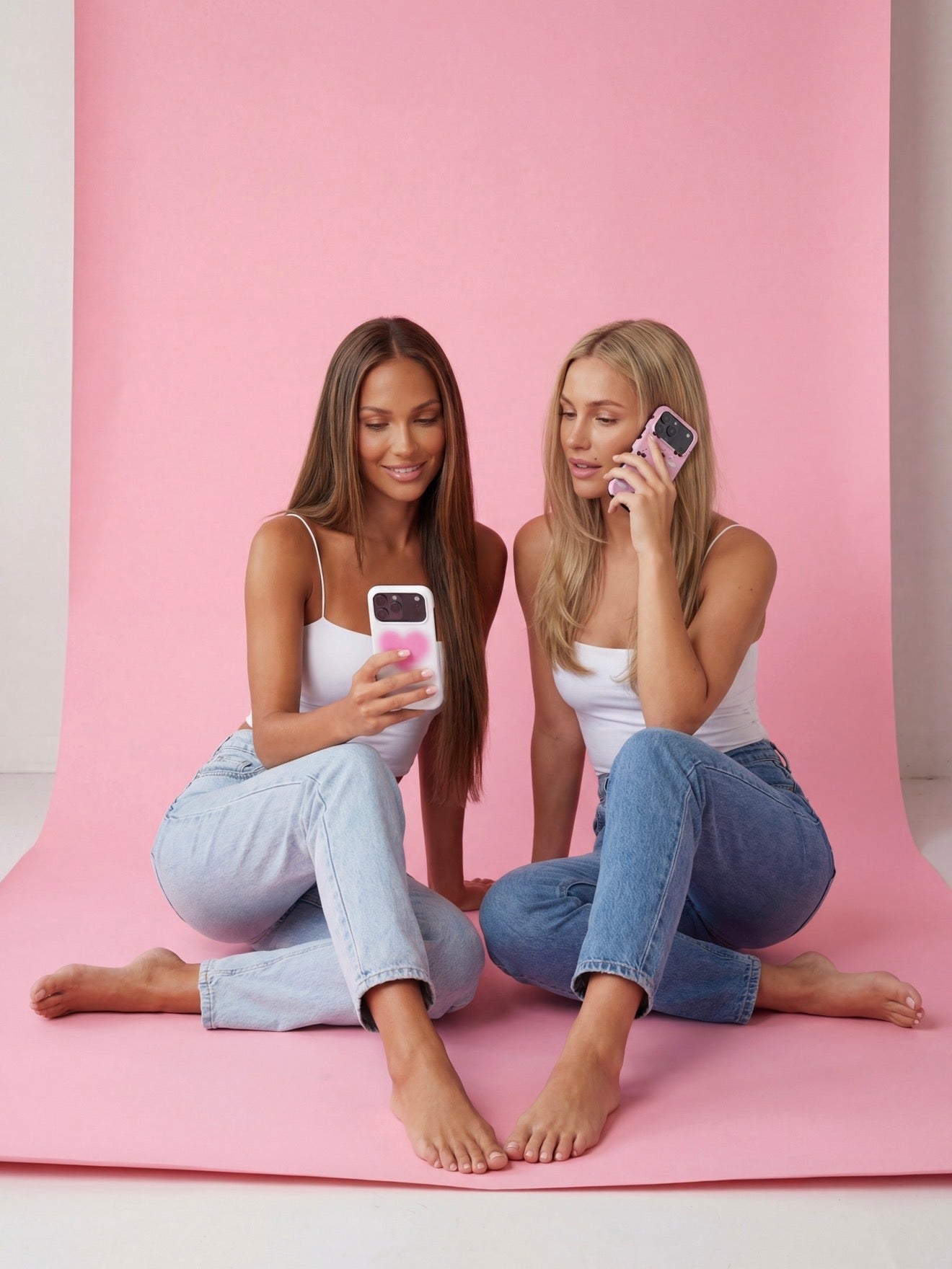 Two stylish women sitting on a pink backdrop, showcasing trendy phone cases with a soft, feminine, and aesthetic vibe.