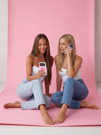Two stylish women sitting on a pink backdrop, showcasing trendy phone cases with a soft, feminine, and aesthetic vibe.