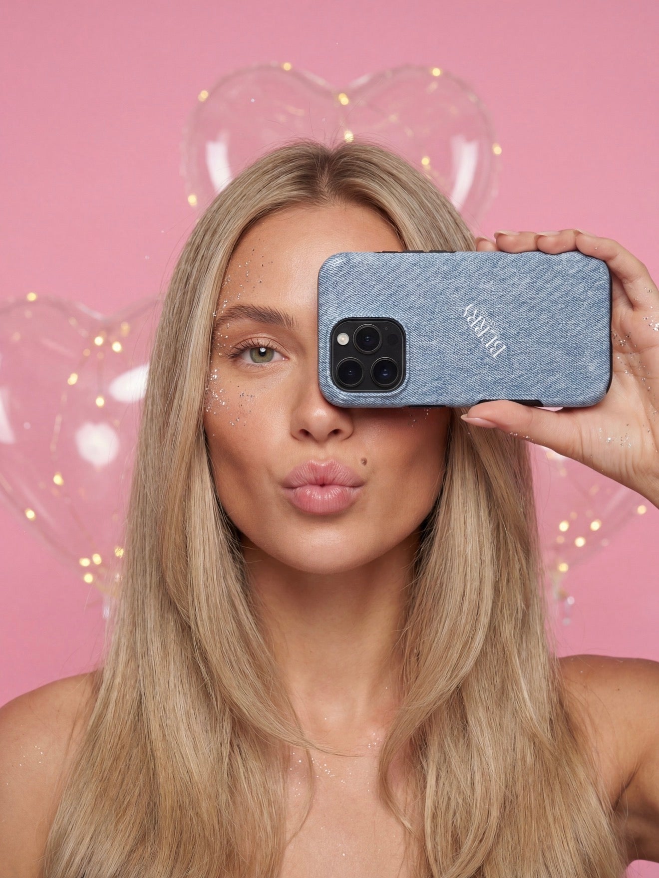 Trendy young woman with long blonde hair holding a blue denim phone case over one eye, posing against a pink background with heart-shaped balloons.