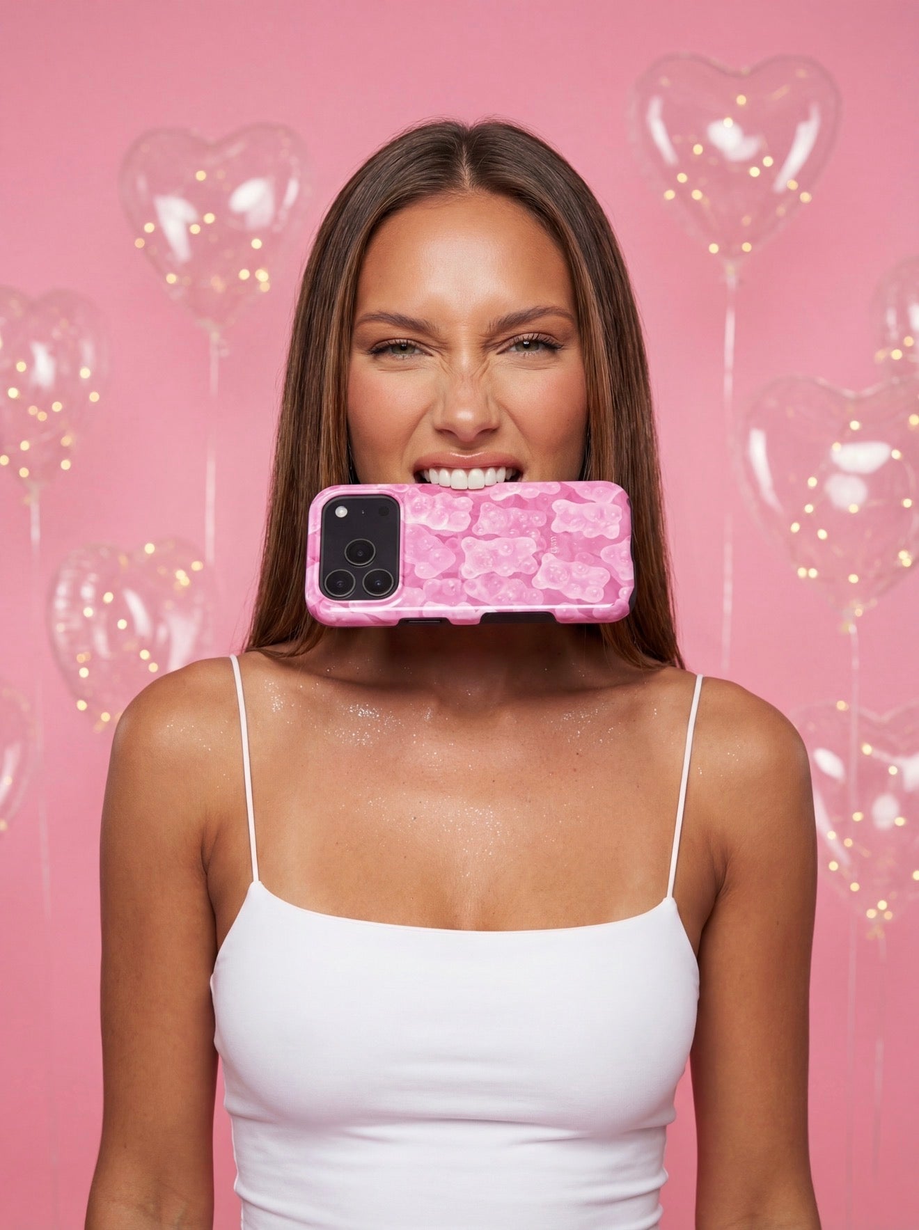 Trendy woman with shiny skin holds a pink gummy-patterned phone case in her mouth against a pink background with heart-shaped balloons.