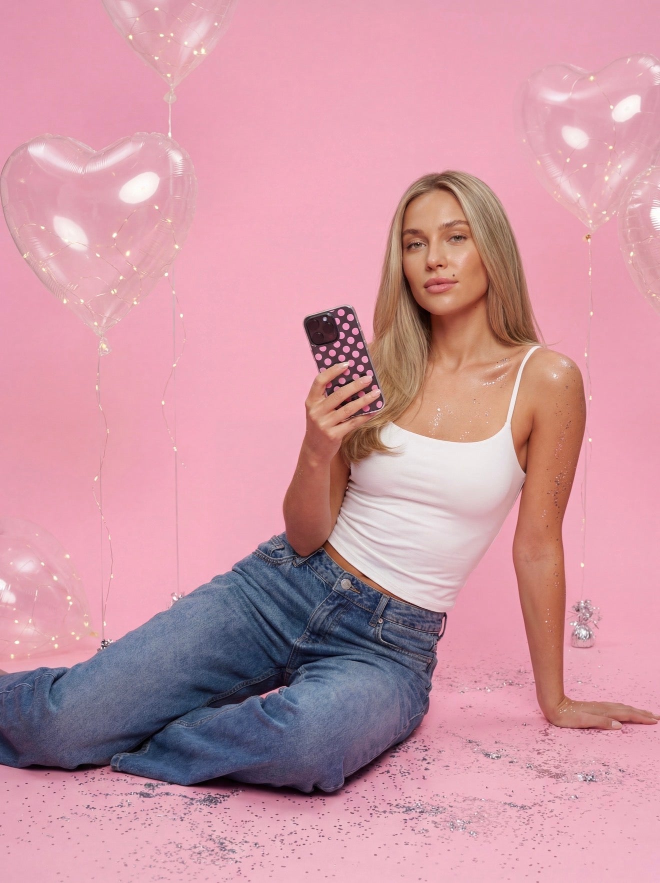 Stylish young woman sitting against pink backdrop holding phone with clear pink polka dot case, surrounded by heart-shaped balloons and glitter for a trendy feminine look.