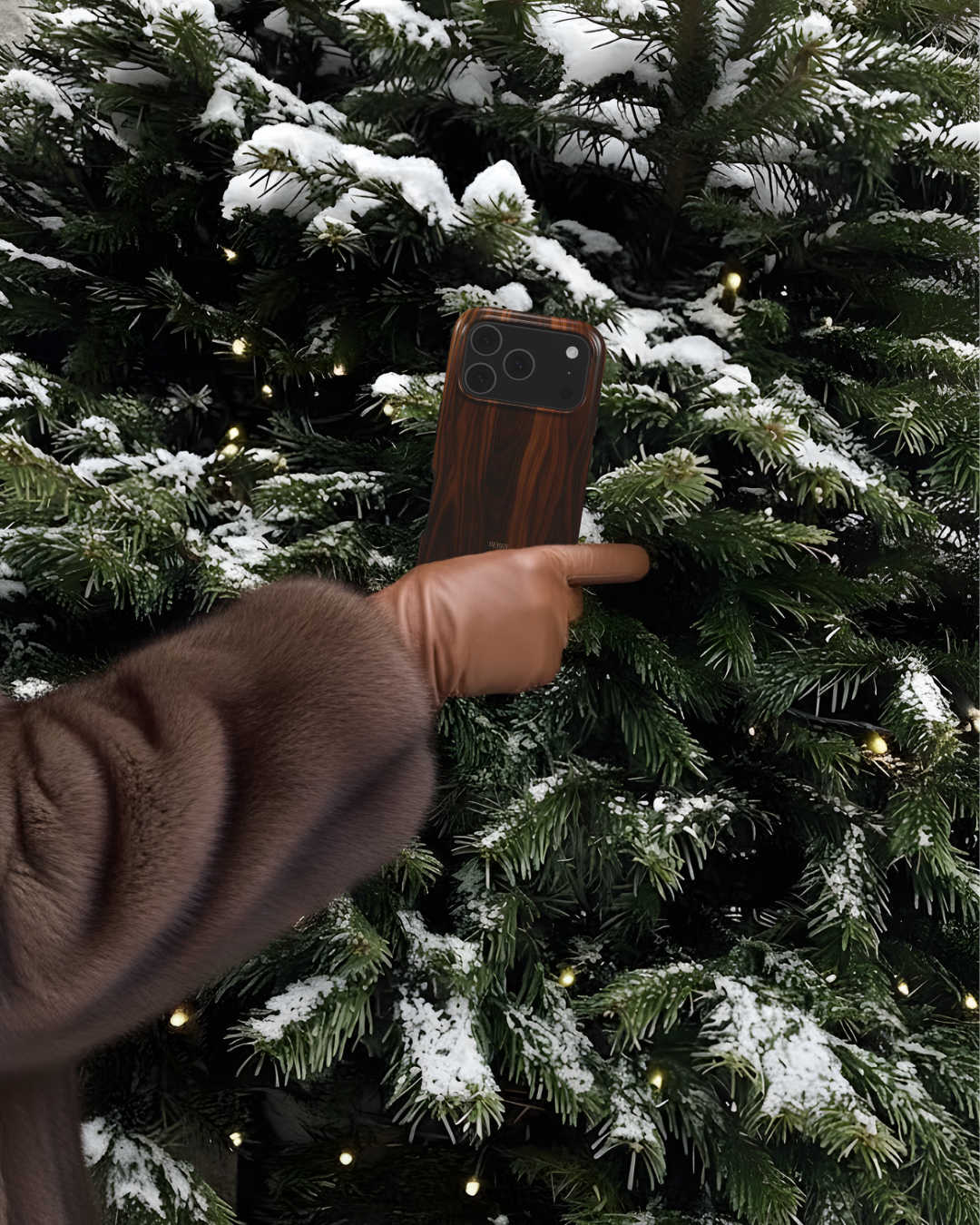 Hand in brown leather glove holding trendy wood-patterned phone case against snow-dusted evergreen branches with twinkling lights in the background.
